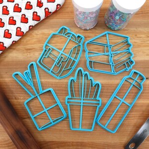 K&ouml;nnte beinhalten: Ein Set aus sechs blauen Plastik-Ausstechern in Form eines Rucksacks, eines Stapels B&uuml;cher, einer Topfpflanze, einer Wasserflasche, einer Geschenkbox und einem Paar Essst&auml;bchen. Die Ausstecher befinden sich auf einer Holzoberfl&auml;che mit einem Nudelholz, einem Spatel und zwei Gl&auml;sern Streusel.