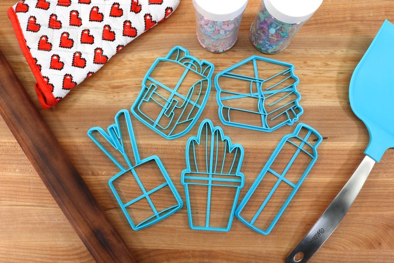 K&ouml;nnte beinhalten: Ein Set aus sechs blauen Plastik-Ausstechern in Form eines Rucksacks, eines Stapels B&uuml;cher, einer Topfpflanze, einer Wasserflasche, einer Geschenkbox und einem Paar Essst&auml;bchen. Die Ausstecher befinden sich auf einer Holzoberfl&auml;che mit einem Nudelholz, einem Spatel und zwei Gl&auml;sern Streusel.