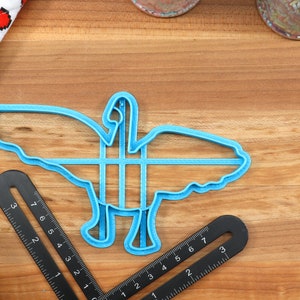 May include: A blue plastic cookie cutter shaped like a goose with its wings spread out. The cutter is on a wooden surface.