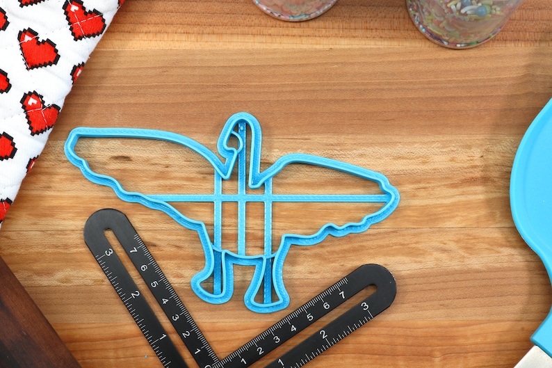 May include: A blue plastic cookie cutter shaped like a goose with its wings spread out. The cutter is on a wooden surface.