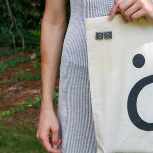 Peut inclure: Un sac fourre-tout en toile blanche avec un point noir et la lettre "&ouml;" imprim&eacute;e dessus. Le sac est orn&eacute; de deux petites &eacute;pingles argent&eacute;es en forme de c&oelig;ur.