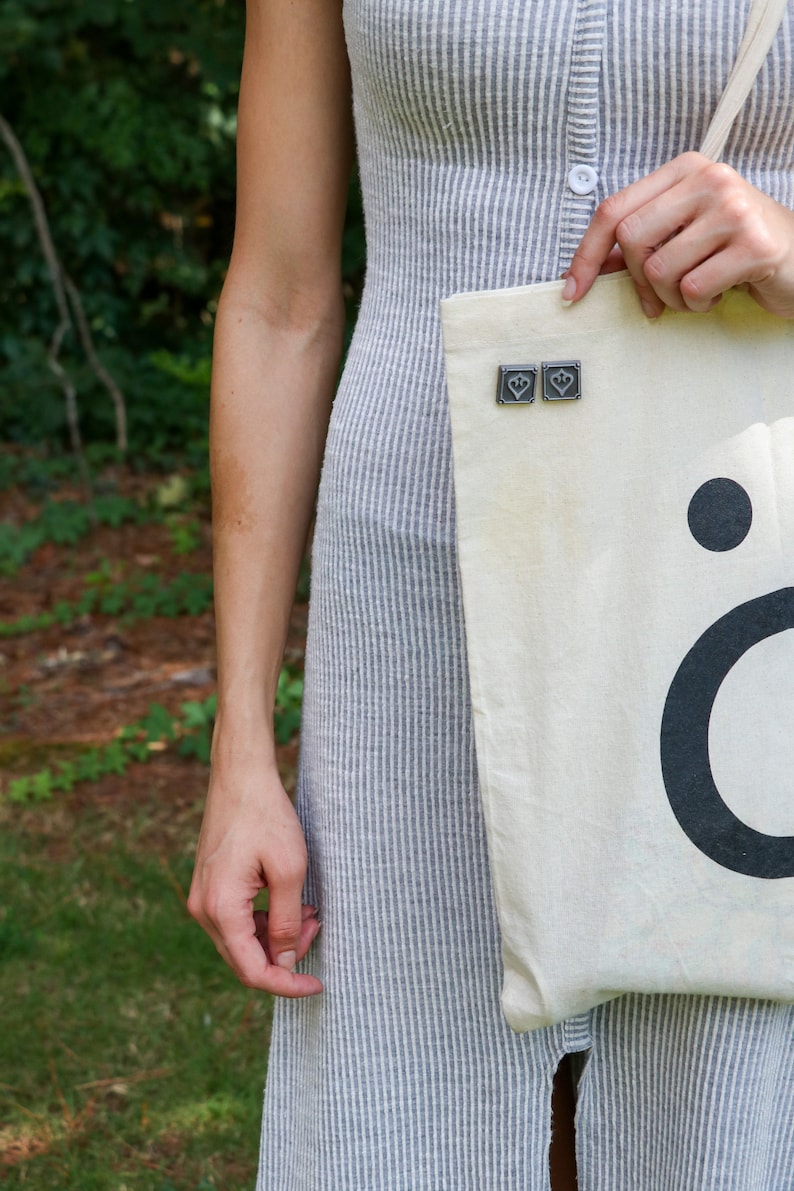 Peut inclure: Un sac fourre-tout en toile blanche avec un point noir et la lettre "&ouml;" imprim&eacute;e dessus. Le sac est orn&eacute; de deux petites &eacute;pingles argent&eacute;es en forme de c&oelig;ur.