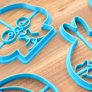 May include: Three blue plastic cookie cutters shaped like a cartoon character with glasses, a bunny rabbit, and a feather. The cutters are on a wooden surface.