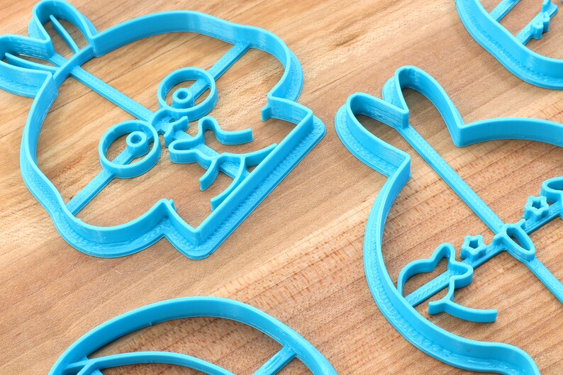 May include: Three blue plastic cookie cutters shaped like a cartoon character with glasses, a bunny rabbit, and a feather. The cutters are on a wooden surface.