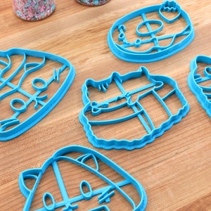 May include: Five blue plastic cookie cutters shaped like cats and a donut. The cutters are on a wooden surface.