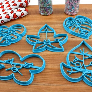 May include: Five blue plastic cookie cutters shaped like leaves. The cutters are on a wooden surface.