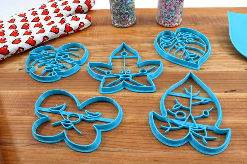 May include: Five blue plastic cookie cutters shaped like leaves. The cutters are on a wooden surface.