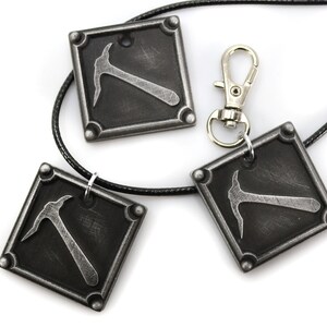 May include: Three square pendants with a pickaxe design, in a distressed silver finish, are displayed on a white background. One pendant is attached to a black cord, and another is connected to a silver clasp. The pendants are likely jewelry.