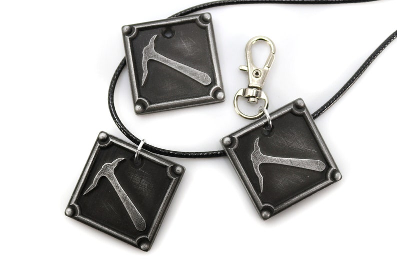 May include: Three square pendants with a pickaxe design, in a distressed silver finish, are displayed on a white background. One pendant is attached to a black cord, and another is connected to a silver clasp. The pendants are likely jewelry.