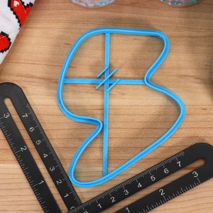 May include: A blue plastic cookie cutter shaped like a heart with a cross-hatch design in the centre. The cutter is on a wooden surface with a ruler.