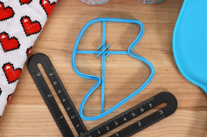 May include: A blue plastic cookie cutter shaped like a heart with a cross-hatch design in the centre. The cutter is on a wooden surface with a ruler.