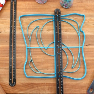 May include: A blue plastic template for making a swirl design on a cake. The template is on a wooden surface with a ruler, rolling pin, and spatula.