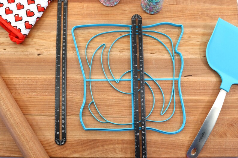 May include: A blue plastic template for making a swirl design on a cake. The template is on a wooden surface with a ruler, rolling pin, and spatula.