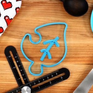May include: A blue plastic cookie cutter shaped like a leaf with a stylized arrow inside. The cutter is on a wooden surface with a ruler and a spatula.