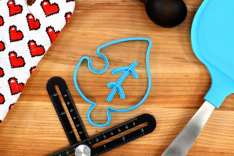 May include: A blue plastic cookie cutter shaped like a leaf with a stylized arrow inside. The cutter is on a wooden surface with a ruler and a spatula.