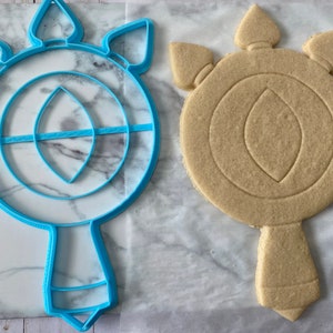 May include: A blue plastic cookie cutter shaped like a stylized eye with a handle. Next to it is a light brown sugar cookie that has been cut out using the cookie cutter.