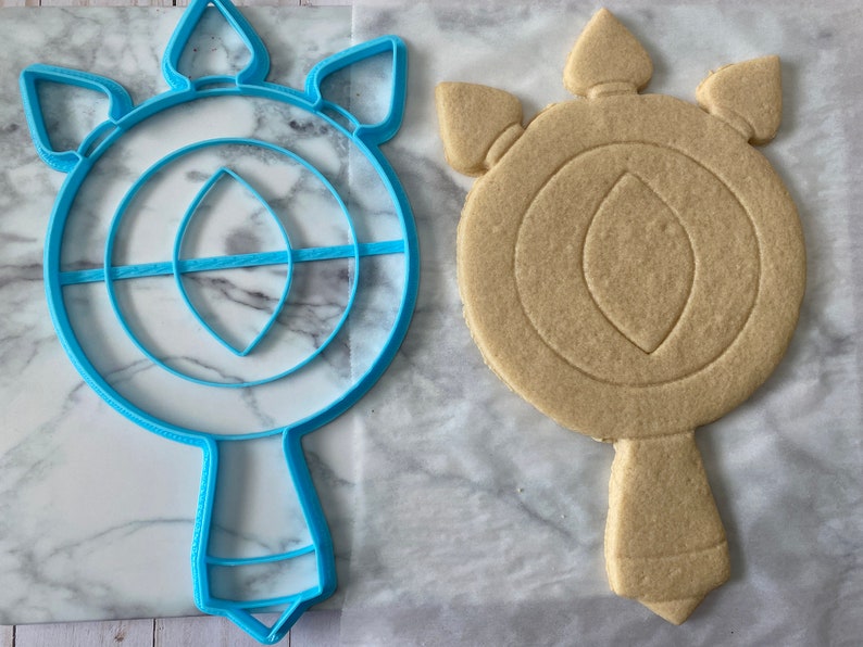 May include: A blue plastic cookie cutter shaped like a stylized eye with a handle. Next to it is a light brown sugar cookie that has been cut out using the cookie cutter.
