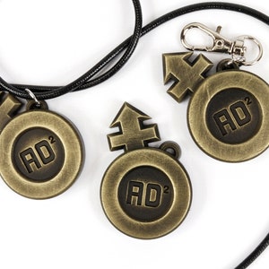 May include: Three gold-colored metal pendants with a black cord. Each pendant features a stylized arrow symbol and the text "AD&sup2;".