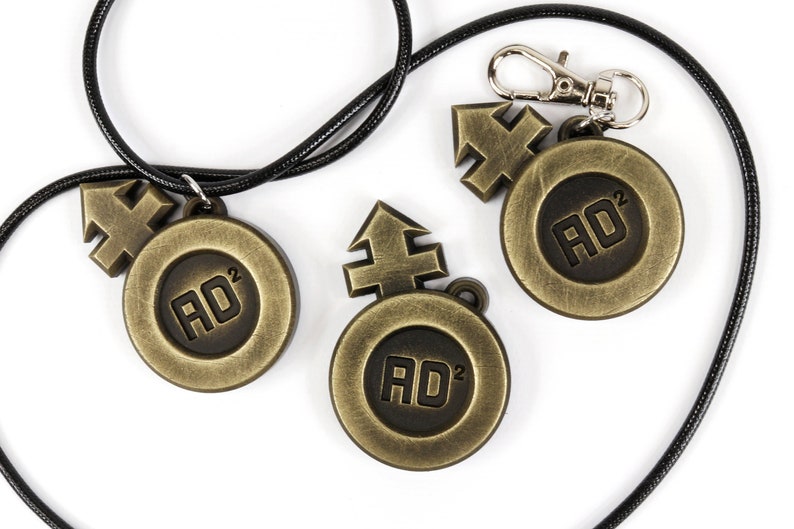 May include: Three gold-colored metal pendants with a black cord. Each pendant features a stylized arrow symbol and the text "AD&sup2;".