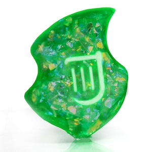 Op de afbeelding: Een groene, iriserende, harsbedel met een wit harp symbool. De bedel is bladvormig en heeft een glinsterende afwerking.