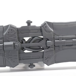 May include: A gray plastic toy weapon with a detailed design, featuring a hinged section and a series of interlocking gears. The toy is made of a durable plastic and has a realistic appearance.