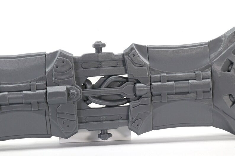 May include: A gray plastic toy weapon with a detailed design, featuring a hinged section and a series of interlocking gears. The toy is made of a durable plastic and has a realistic appearance.
