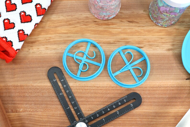 May include: Two blue plastic cookie cutters shaped like the letter "S" with a decorative flourish. The cutters are on a wooden surface.
