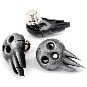 Op de afbeelding: Drie zilverkleurige schedelspelden met een textuur. Elke speld heeft een schedel met drie gaten en een vleugelachtig ontwerp. De spelden hebben een zilverkleurige metalen achterkant met een speldmechanisme.