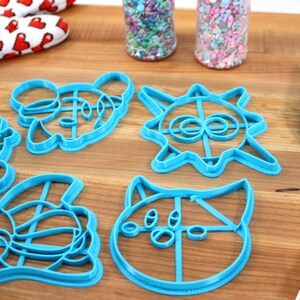 May include: Five blue plastic cookie cutters shaped like cartoon characters on a wooden surface. The cutters are in the shape of a cat, a bear, a sun, a fox, and a panda.