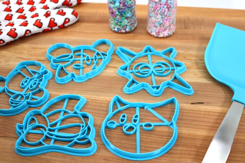 May include: Five blue plastic cookie cutters shaped like cartoon characters on a wooden surface. The cutters are in the shape of a cat, a bear, a sun, a fox, and a panda.