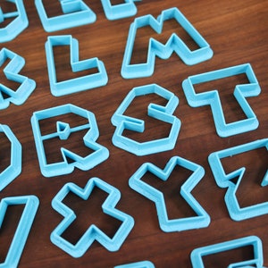 May include: A set of blue plastic cookie cutters shaped like letters of the alphabet. The cutters are arranged on a wooden surface.