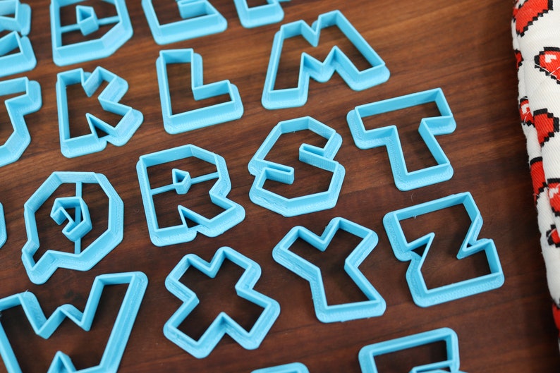 May include: A set of blue plastic cookie cutters shaped like letters of the alphabet. The cutters are arranged on a wooden surface.