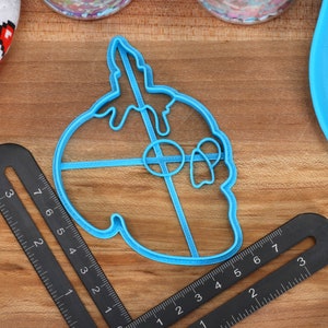 May include: A blue plastic cookie cutter shaped like a skull with dripping icing. The cutter is on a wooden surface with a ruler and a blue plate.