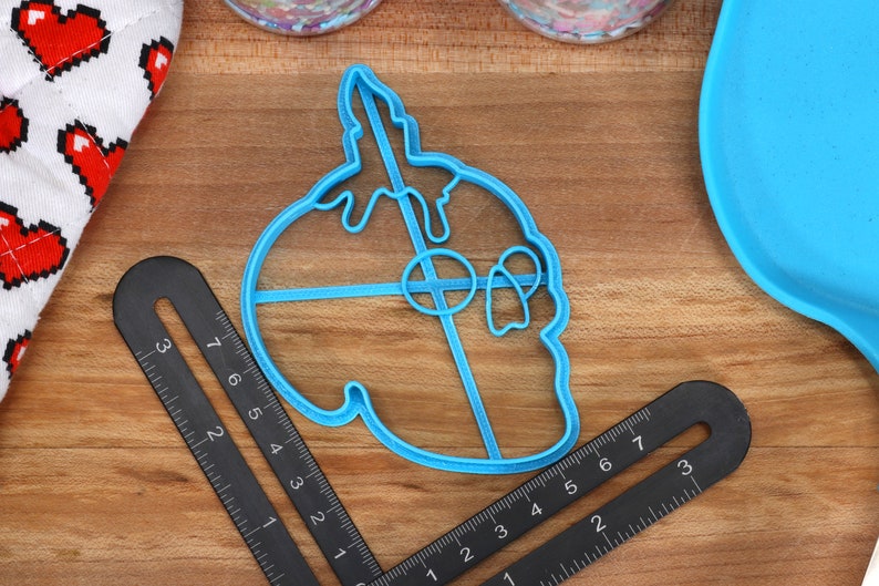 May include: A blue plastic cookie cutter shaped like a skull with dripping icing. The cutter is on a wooden surface with a ruler and a blue plate.