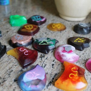 May include: A collection of colorful resin dice with various symbols and numbers. The dice are in different shapes and sizes, and some have a marbled effect.