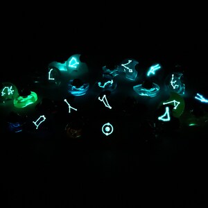May include: A collection of glowing dice with constellations printed on each face. The dice are a variety of colours, including green, blue, and purple. The constellations are visible in the dark.