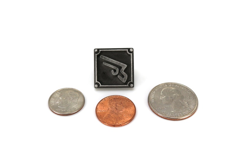 May include: A silver metal pin with a black square background and a silver stylized wing design. The pin is surrounded by three US coins: a quarter, a dime, and a penny.