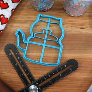 May include: A blue plastic cookie cutter shaped like a teapot with a handle and spout. The cutter is on a wooden surface with a ruler and a blue spatula.