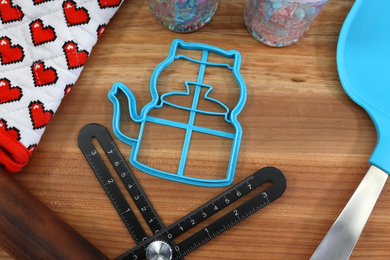 May include: A blue plastic cookie cutter shaped like a teapot with a handle and spout. The cutter is on a wooden surface with a ruler and a blue spatula.