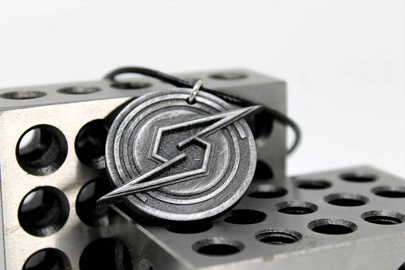 May include: A silver-colored pendant necklace featuring a stylized 'S' symbol with a lightning bolt design. The pendant is circular and has a textured surface. The necklace is displayed on a gray metal surface.