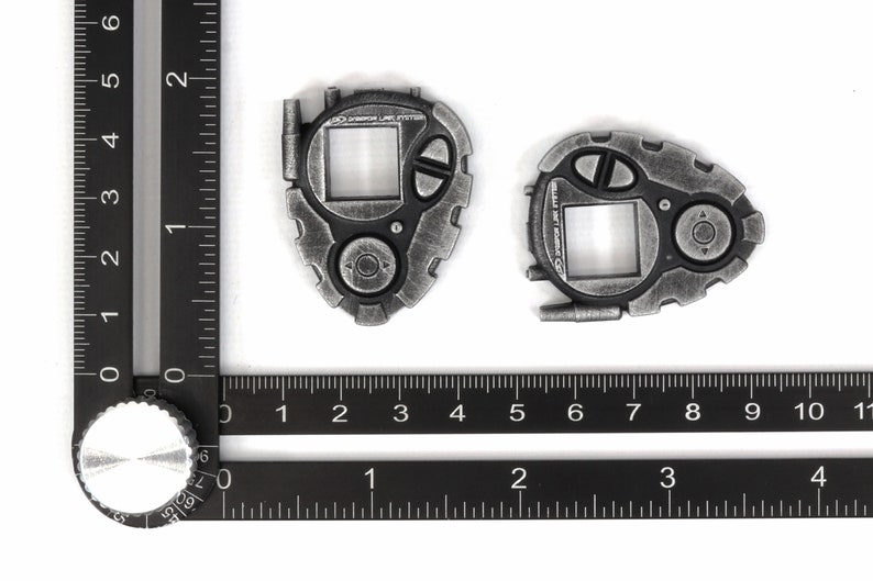May include: Two silver-colored Digimon-style digital pet devices with a screen, buttons, and a small antenna. The devices are on a black ruler with white markings.