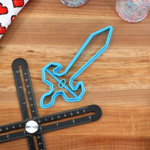 May include: A blue plastic cookie cutter shaped like a sword. The cutter is on a wooden surface with a ruler and other baking supplies.