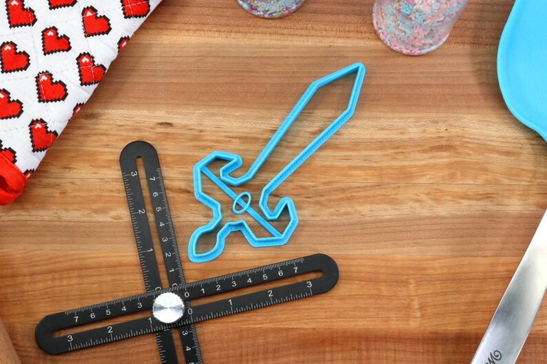 May include: A blue plastic cookie cutter shaped like a sword. The cutter is on a wooden surface with a ruler and other baking supplies.