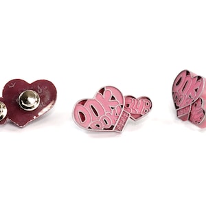 May include: Three pink heart-shaped pins with the text "Book Club" on them. The pins have a silver metal backing and a safety pin closure.