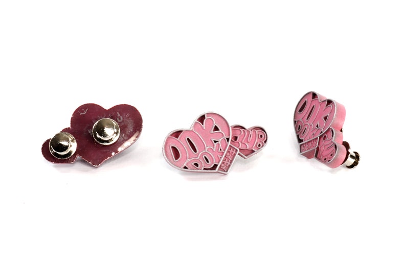 May include: Three pink heart-shaped pins with the text "Book Club" on them. The pins have a silver metal backing and a safety pin closure.