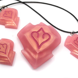 May include: Pink resin pendants and charms with a heart-shaped design. One pendant is on a black cord necklace, another has a silver clasp. The other two charms are loose. The pendants and charms have a glittery, iridescent finish.