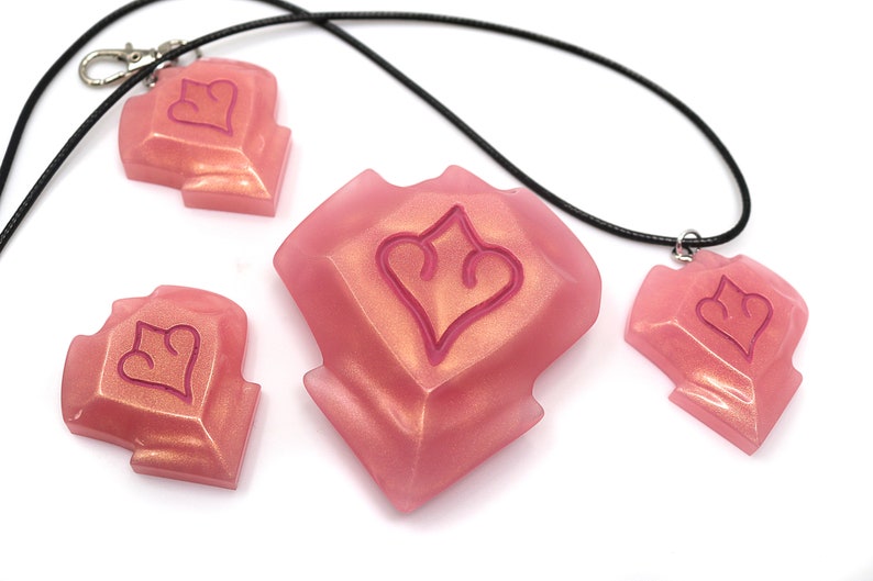 May include: Pink resin pendants and charms with a heart-shaped design. One pendant is on a black cord necklace, another has a silver clasp. The other two charms are loose. The pendants and charms have a glittery, iridescent finish.