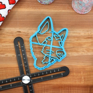 May include: A blue plastic cookie cutter shaped like a dog's head. The cutter is on a wooden surface with a ruler and a spatula.
