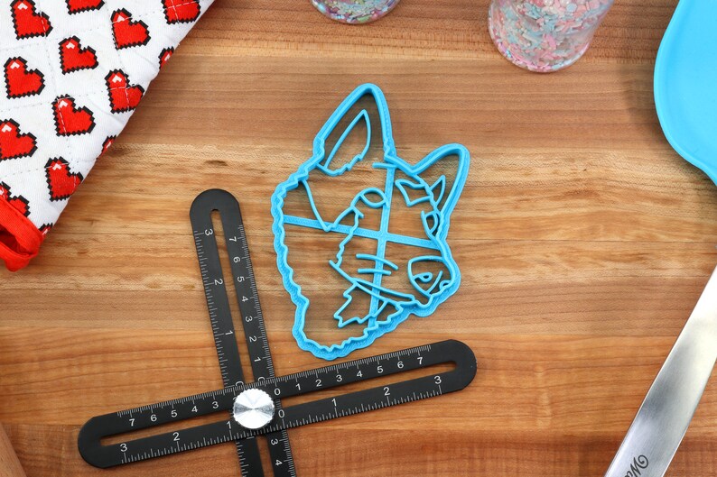 May include: A blue plastic cookie cutter shaped like a dog's head. The cutter is on a wooden surface with a ruler and a spatula.