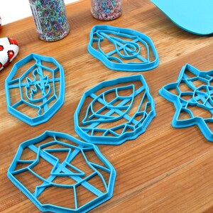 May include: Five blue plastic cookie cutters on a wooden surface. The cutters are shaped like a leaf, a star, a geometric design, and two different geometric shapes.